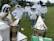 A group of people wearing protective beekeeping suits with veils stand in a grassy field. Two metal bee smokers are in the foreground, emitting smoke. The individuals are dressed uniformly, with white suits and green gloves, and trees are visible in the background.