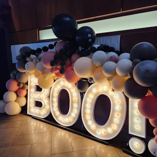 A decorative display with large illuminated letters spelling 'BOO!' surrounded by numerous balloons in various colors. The balloons include shades of black, gray, white, yellow, peach, and pink, with some featuring googly eyes. This appears to be a festive arrangement set against a dark background with lit floor tiles.
