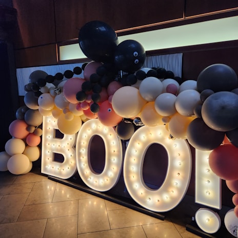 A decorative display with large illuminated letters spelling 'BOO!' surrounded by numerous balloons in various colors. The balloons include shades of black, gray, white, yellow, peach, and pink, with some featuring googly eyes. This appears to be a festive arrangement set against a dark background with lit floor tiles.
