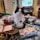 A black and white cat sits amidst various colorful wrapped gifts and decorative items on a table. A television in the background displays the text 'Crispy Garlic'. The room contains assorted furniture and decorations, adding to a cozy, cluttered atmosphere.