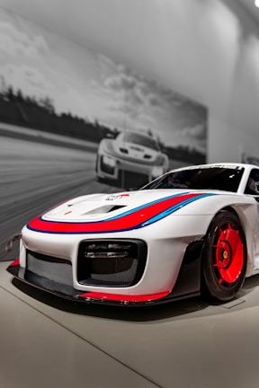 A sleek, high-performance sports car with a white body and striking red and blue accents is prominently displayed in the foreground. Its aerodynamic design features large vents and a low front profile. In the background, a black-and-white image of a similar car racing on a track is visible, adding a dynamic and energetic atmosphere.