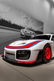A sleek, high-performance sports car with a white body and striking red and blue accents is prominently displayed in the foreground. Its aerodynamic design features large vents and a low front profile. In the background, a black-and-white image of a similar car racing on a track is visible, adding a dynamic and energetic atmosphere.