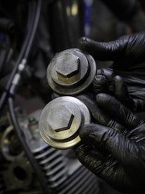 A pair of black gloved hands holding two metallic hexagonal caps, likely engine or machinery parts. The background shows part of a mechanical engine, indicating a workshop setting.