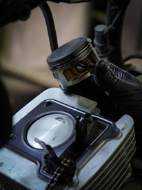 A hand wearing a black glove holds a metallic piston above an open engine cylinder. The engine components are detailed, with visible bolts and a smooth metal surface inside the cylinder.
