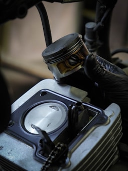 A hand wearing a black glove holds a metallic piston above an open engine cylinder. The engine components are detailed, with visible bolts and a smooth metal surface inside the cylinder.