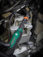 A close-up view of a hand wearing a glove holding a torque wrench near the engine components of a motorcycle or vehicle. The wrench features a green and black handle with a visible measurement scale, and the background shows metallic engine parts and cables.