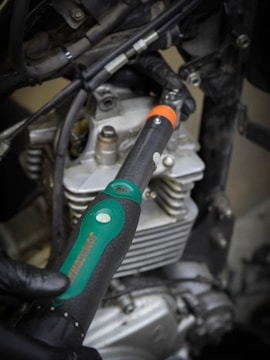 A close-up view of a hand wearing a glove holding a torque wrench near the engine components of a motorcycle or vehicle. The wrench features a green and black handle with a visible measurement scale, and the background shows metallic engine parts and cables.