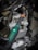 A close-up view of a hand wearing a glove holding a torque wrench near the engine components of a motorcycle or vehicle. The wrench features a green and black handle with a visible measurement scale, and the background shows metallic engine parts and cables.