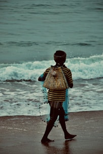 A woman walking along the shore in a flowy sundress, holding a colorful beachcraft tote bag.