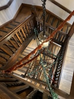 A multi-story wooden staircase adorned with festive green and orange garlands. The staircase is spiraling downwards with a classic and ornate railing, and a large vintage-style chandelier hangs in the center. The wood has a rich, dark color and there is a blend of decorative and elegant details.
