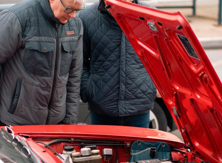 two men looking under the hood of a red car