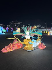 A vibrant and colorful outdoor display featuring illuminated sculptures. The centerpiece is a large artistic rendition of a mythical creature with a human face and wings extending outward, surrounded by abstract forms and elements. Brightly colored lights enhance the intricate designs, contrasting against the dark night backdrop. Various other lit sculptures can be seen scattered in the background, adding to the festive atmosphere.