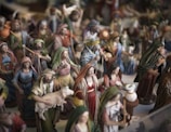 A collection of painted figurines depicting various people in a traditional setting. These figures are dressed in robes and cloaks, some carrying items such as baskets or animals. A notable figure is holding a sheep, and another appears to be pouring or holding something near a white container.