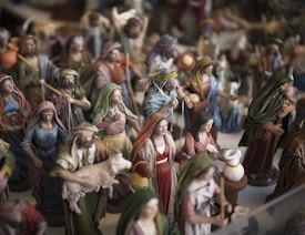 A collection of painted figurines depicting various people in a traditional setting. These figures are dressed in robes and cloaks, some carrying items such as baskets or animals. A notable figure is holding a sheep, and another appears to be pouring or holding something near a white container.