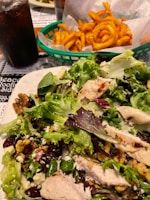 A fresh salad with grilled chicken slices, walnuts, leafy greens, and possibly cranberries is served on a plate. Nearby, a basket lined with paper contains seasoned curly fries. A cold beverage, likely soda, is placed in a glass on the left side.