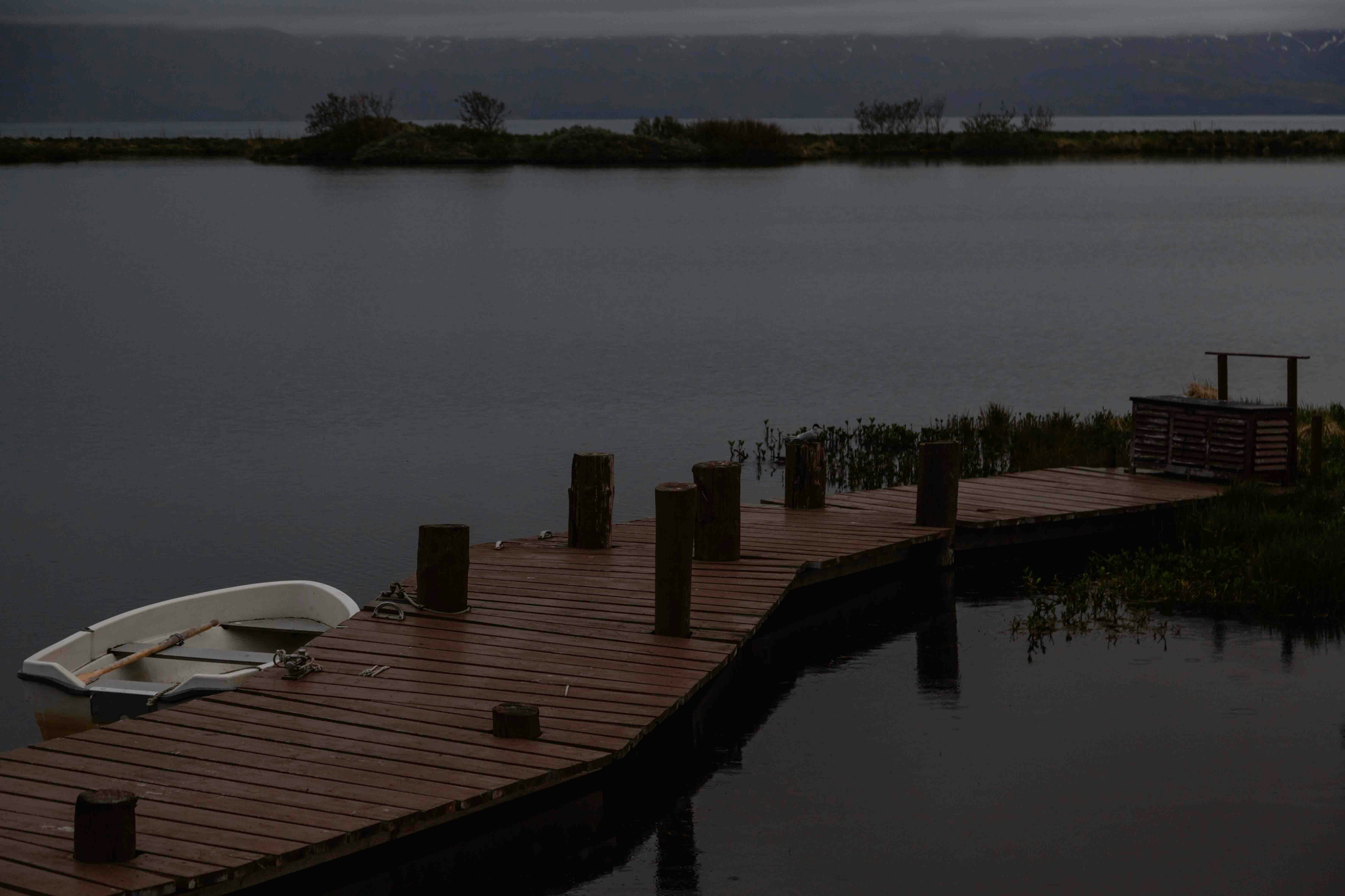 A serene lakeside scene featuring a wooden dock with a single rowboat, a bench, and a view of distant mountains under an overcast sky.