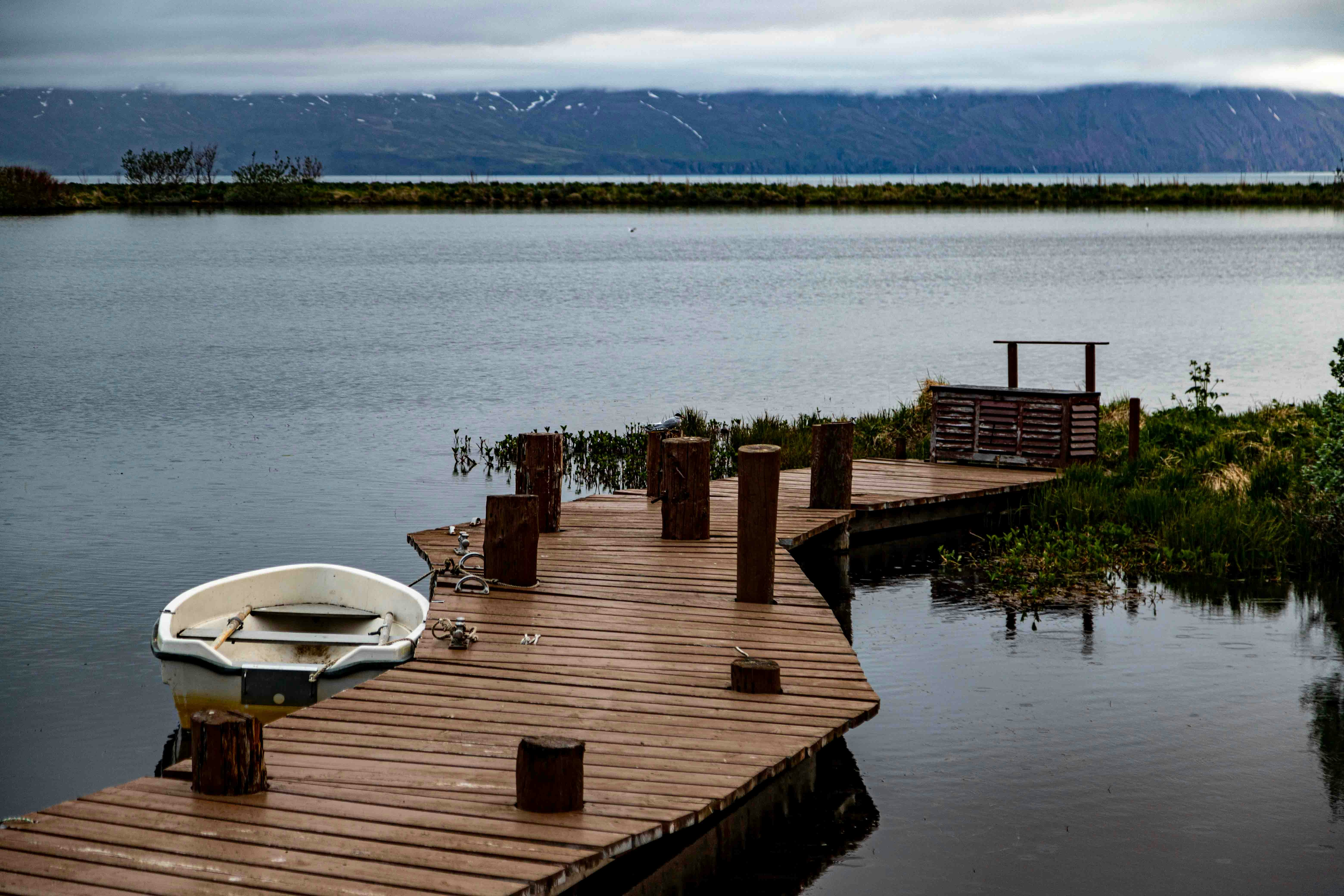 a boat is docked on a wooden dock, A serene lakeside scene featuring a wooden dock with a single rowboat, a bench, and a view of distant mountains under an overcast sky.