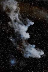 Wispy nebula with a ghostly shape set against a backdrop of countless stars. The nebula features cloud-like structures with varying intensities of brightness, surrounded by tiny specks of light representing distant stars.