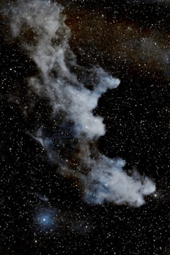 Wispy nebula with a ghostly shape set against a backdrop of countless stars. The nebula features cloud-like structures with varying intensities of brightness, surrounded by tiny specks of light representing distant stars.
