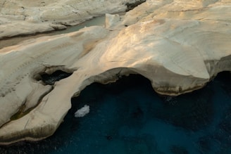 A landscape featuring large, smooth sandstone formations with light beige hues. The rock structures appear carved by natural processes, creating arches and overhangs. Below, the deep blue of the ocean contrasts with the pale rocks, highlighting their texture and shapes. The scene is devoid of people, emphasizing the natural, untouched quality of the environment.