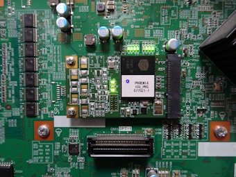 A printed circuit board with various electronic components and integrated circuits, prominently featuring a large black chip labeled 'PHOENIX ICU PRG 0770Z1-1'. Several capacitors are visible, along with numerous small resistors and connectors. There is a green LED light illuminated on the board, and the surface is covered with intricate traces and solder points.