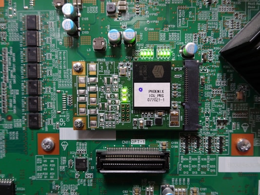 A printed circuit board with various electronic components and integrated circuits, prominently featuring a large black chip labeled 'PHOENIX ICU PRG 0770Z1-1'. Several capacitors are visible, along with numerous small resistors and connectors. There is a green LED light illuminated on the board, and the surface is covered with intricate traces and solder points.
