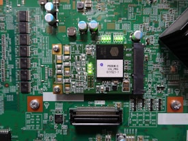 A printed circuit board with various electronic components and integrated circuits, prominently featuring a large black chip labeled 'PHOENIX ICU PRG 0770Z1-1'. Several capacitors are visible, along with numerous small resistors and connectors. There is a green LED light illuminated on the board, and the surface is covered with intricate traces and solder points.