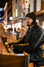 A person wearing a beanie and glasses, playing a wooden piano on a busy street at night. The scene is illuminated by the warm glow of streetlights and store signs, with a blurred background of people walking.