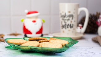 A festive scene features a green star-shaped plate with several round cookies placed on it. In the background, there's a white mug with a Christmas-themed design and a ceramic decoration of Santa Claus. The setting appears to be a kitchen counter with a marble-like surface.