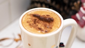 A creamy cup of coffee topped with frothy milk and a dusting of cocoa powder, placed in a white mug. The background is slightly blurred, featuring hints of festive decor and possibly pine cones.