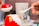 A hand is holding a small spoon filled with sugar and is placing it into a red and white decorative jar, which is open at the top. The jar resembles a Santa Claus figure with its red and white colors. In the background, there is a blurred tall cup with text on it.
