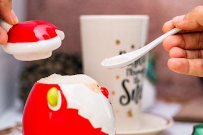 A hand is holding a small spoon filled with sugar and is placing it into a red and white decorative jar, which is open at the top. The jar resembles a Santa Claus figure with its red and white colors. In the background, there is a blurred tall cup with text on it.