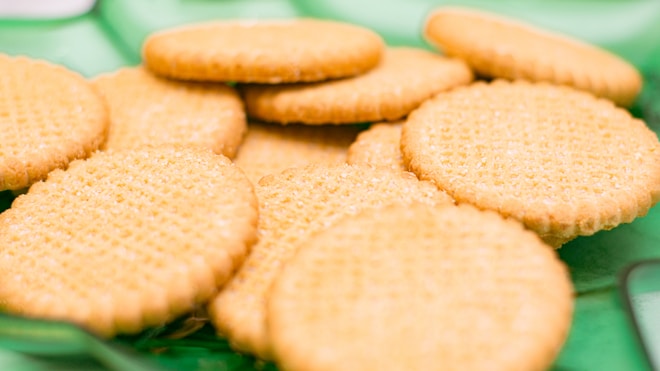 A pile of round, golden brown biscuits with a textured surface is arranged on a green background. The biscuits appear crisp with a slight sheen on their surface.