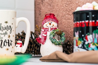a snowman holding a wreath next to two coffee mugs