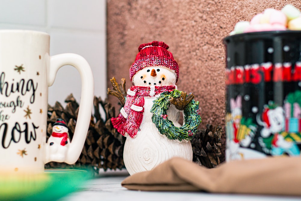 a snowman holding a wreath next to two coffee mugs