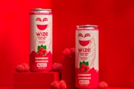 Two cans of Wize Sparkling Iced Tea Raspberry flavor are placed on a red surface with several fresh raspberries around the cans. The cans are primarily white and red with a smiling face and the words 'Wize' and 'Raspberry Framboise' prominently displayed. The background is entirely red, creating a vibrant and eye-catching scene.