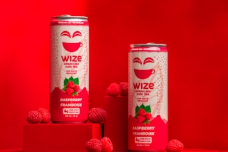 Two cans of Wize Sparkling Iced Tea Raspberry flavor are placed on a red surface with several fresh raspberries around the cans. The cans are primarily white and red with a smiling face and the words 'Wize' and 'Raspberry Framboise' prominently displayed. The background is entirely red, creating a vibrant and eye-catching scene.