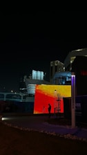 A nighttime urban scene featuring modern architecture with illuminated buildings. A large, vibrant LED screen displays an abstract red and orange pattern. A person is silhouetted in front of the screen, standing near a tripod, suggesting photography or video recording activities. Surrounding structures are dimly lit, creating a contrast with the bright lights of the screen.