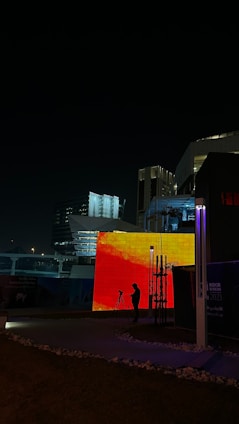 A nighttime urban scene featuring modern architecture with illuminated buildings. A large, vibrant LED screen displays an abstract red and orange pattern. A person is silhouetted in front of the screen, standing near a tripod, suggesting photography or video recording activities. Surrounding structures are dimly lit, creating a contrast with the bright lights of the screen.