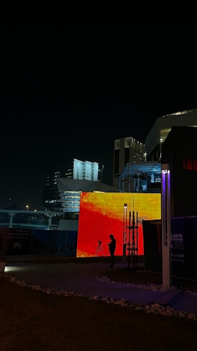 A nighttime urban scene featuring modern architecture with illuminated buildings. A large, vibrant LED screen displays an abstract red and orange pattern. A person is silhouetted in front of the screen, standing near a tripod, suggesting photography or video recording activities. Surrounding structures are dimly lit, creating a contrast with the bright lights of the screen.