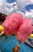 Two large, fluffy pink cotton candy sticks are held in front of a bright blue sky with scattered clouds. A hand is visible holding the sticks over a shallow pool of water. In the background, there are people gathered, some wearing red shirts, suggesting an outdoor event or fair setting.