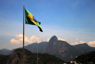 A Brazilian flag waves in the foreground with a mountainous landscape in the background. The iconic Christ the Redeemer statue is visible atop one of the mountains under a clear blue sky.