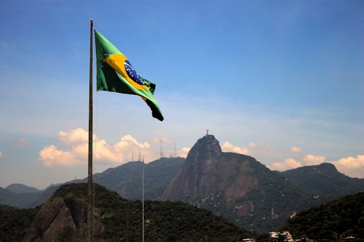 A Brazilian flag waves in the foreground with a mountainous landscape in the background. The iconic Christ the Redeemer statue is visible atop one of the mountains under a clear blue sky.