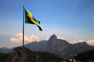A Brazilian flag waves in the foreground with a mountainous landscape in the background. The iconic Christ the Redeemer statue is visible atop one of the mountains under a clear blue sky.
