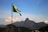 A Brazilian flag waves in the foreground with a mountainous landscape in the background. The iconic Christ the Redeemer statue is visible atop one of the mountains under a clear blue sky.