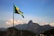A Brazilian flag waves in the foreground with a mountainous landscape in the background. The iconic Christ the Redeemer statue is visible atop one of the mountains under a clear blue sky.