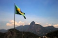 A Brazilian flag waves in the foreground with a mountainous landscape in the background. The iconic Christ the Redeemer statue is visible atop one of the mountains under a clear blue sky.
