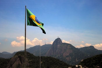 A Brazilian flag waves in the foreground with a mountainous landscape in the background. The iconic Christ the Redeemer statue is visible atop one of the mountains under a clear blue sky.