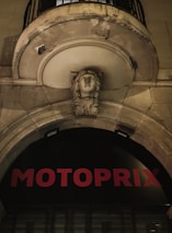 An architectural detail features a stone arch with a sculpted face in the center. Above it, there is a balcony with a metal railing. The facade has signs of weathering and wear. Below the arch, red letters spell out 'MOTOPRIX' against a black background.
