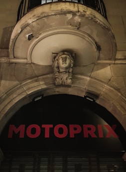 An architectural detail features a stone arch with a sculpted face in the center. Above it, there is a balcony with a metal railing. The facade has signs of weathering and wear. Below the arch, red letters spell out 'MOTOPRIX' against a black background.